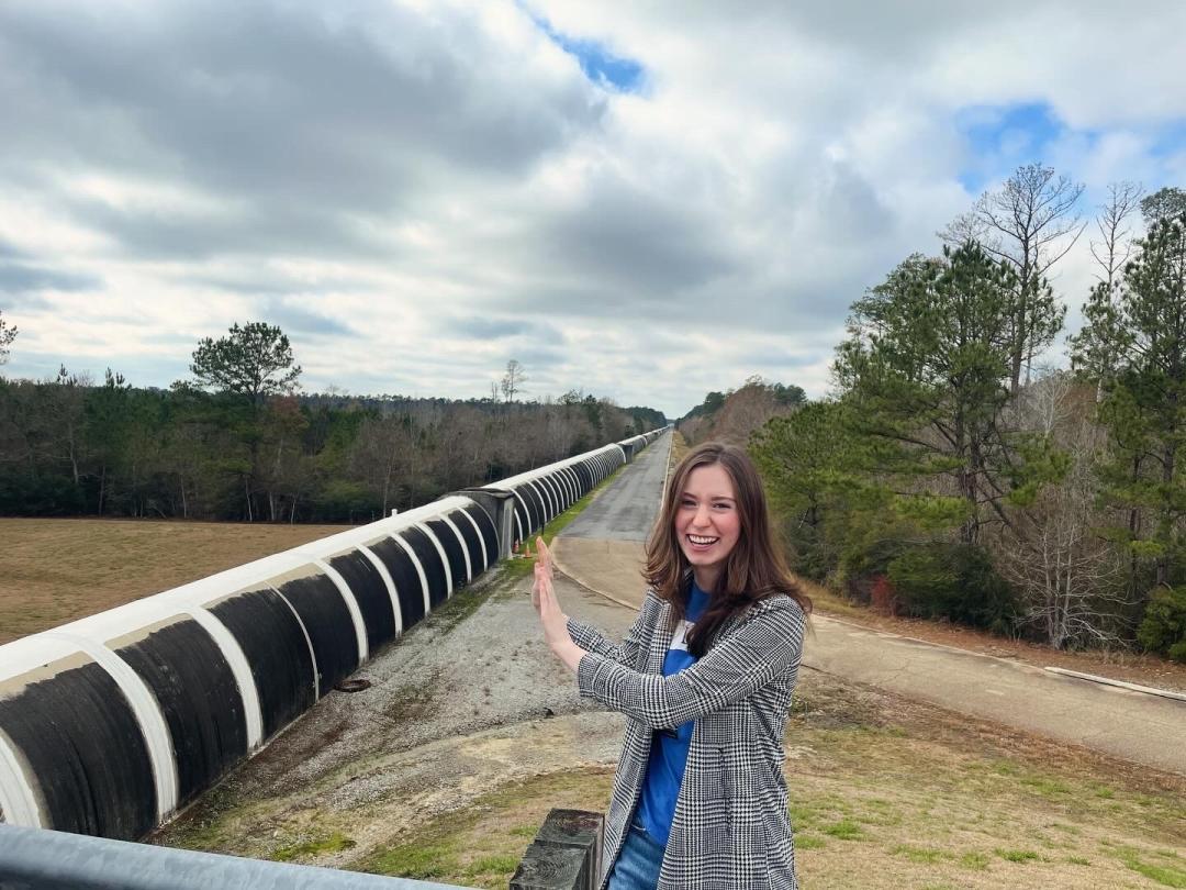 A long, tunnel-like structure stretches to the horizon amidst trees and a dirt road. In the foreground a woman is smiling with her hands pointing to the structure.