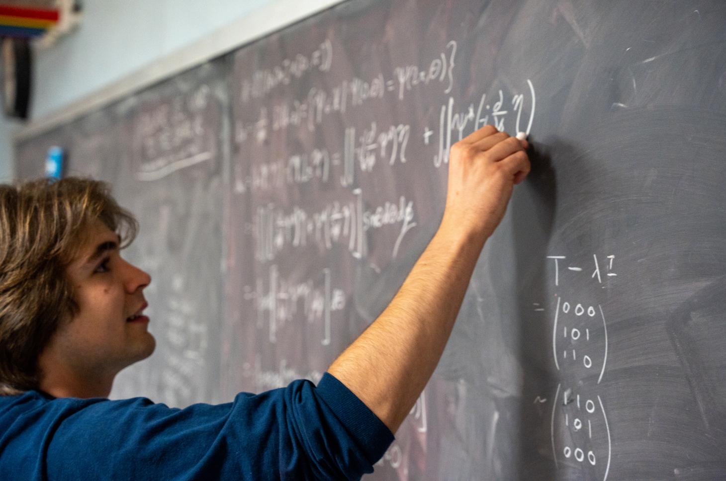 Joey Takach grinning while writing equations on a chalkboard.
