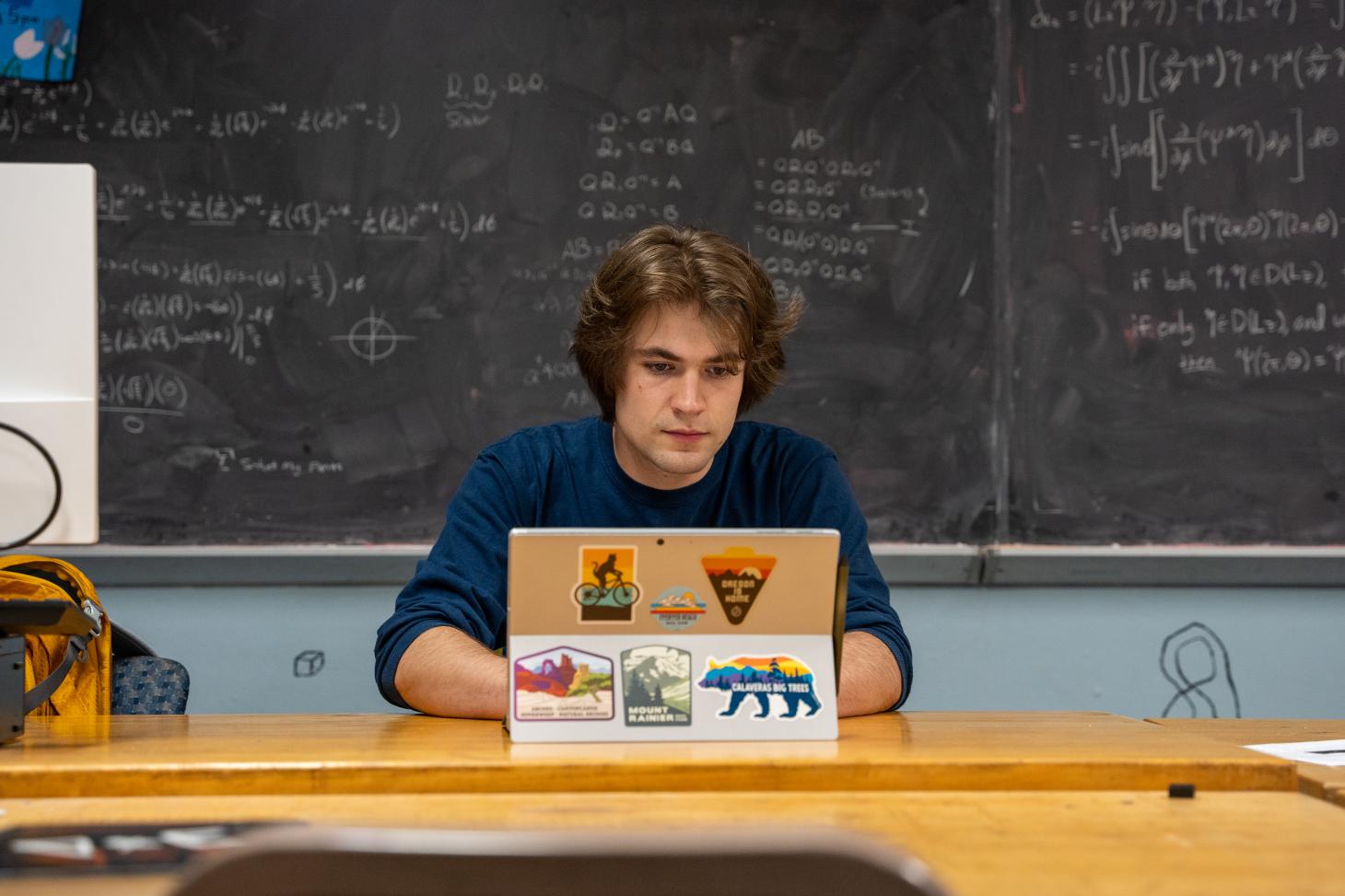 Takach is busy typing behind his laptop. A chalkboard full of mathematical equations stands behind him.