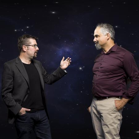 Two men stand in front of a background of space on a green screen.