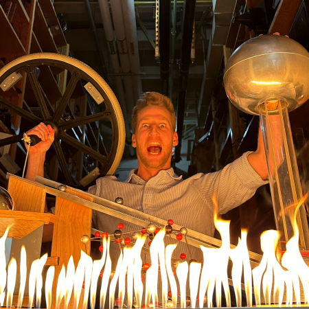 A man in a stripped shirt holds a bicycle wheel and other physics demonstration tools sit on the table in front of him and there is fire at the bottom of the photo.