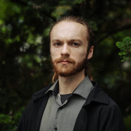 A man in a grey shirt and black coat poses for a photo in front of trees.