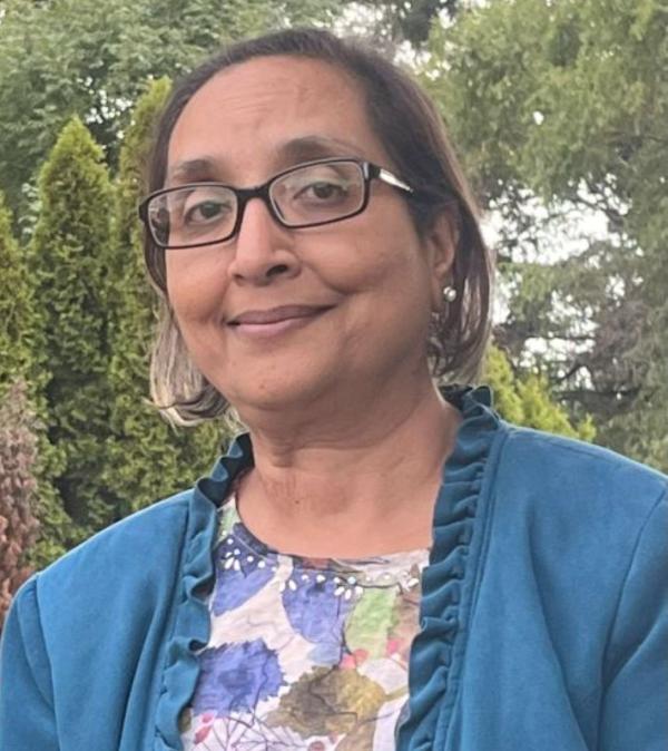 Person wearing glasses and a blue jacket over a floral-patterned top, standing outdoors in front of dense green trees and shrubs in a garden-like setting.